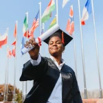 Formanda sorrindo com capelo e beca, erguendo seu diploma em frente a diversas bandeiras internacionais, simbolizando o sucesso e as oportunidades de fazer uma faculdade no exterior.