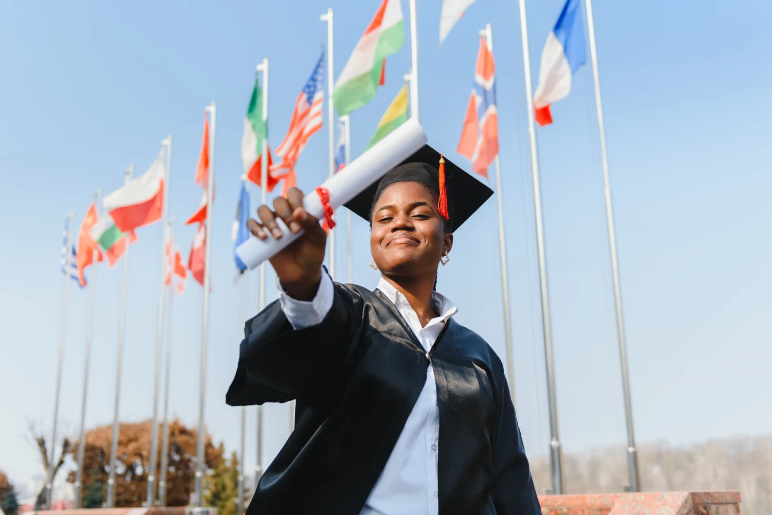 Formanda sorrindo com capelo e beca, erguendo seu diploma em frente a diversas bandeiras internacionais, simbolizando o sucesso e as oportunidades de fazer uma faculdade no exterior.